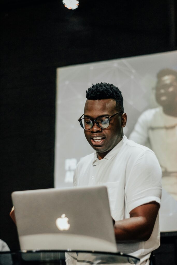 A man in glasses smiles while using an Apple laptop during virtual events.